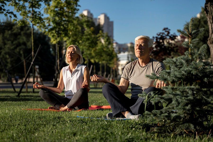 Two people sit in a cross-legged meditative pose on yoga mats in a grassy park, eyes closed and arms extended.