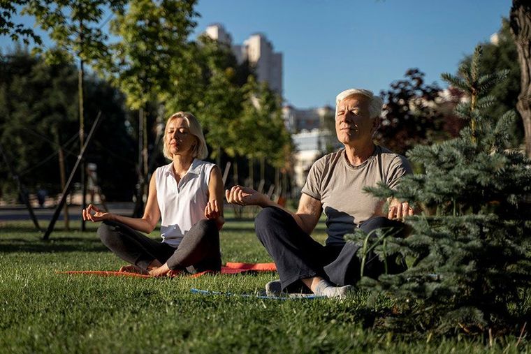 Two people sit in a cross-legged meditative pose on yoga mats in a grassy park, eyes closed and arms extended.