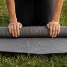 A person kneeling on the grass rolls up a textured gray exercise mat.