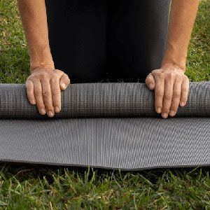 A person kneeling on the grass rolls up a textured gray exercise mat.