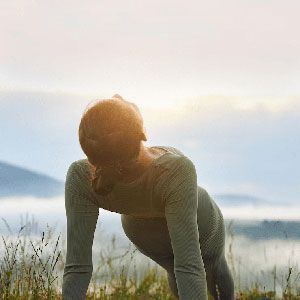 A person in workout attire performs a plank pose in a grassy field during sunrise with misty mountains in the background.