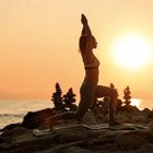 A person performs a yoga lunge with arms raised against a sunset over a rocky coastline.