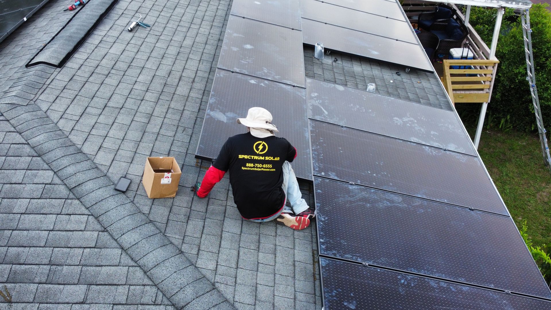 A man is sitting on the roof of a house working on solar panels.