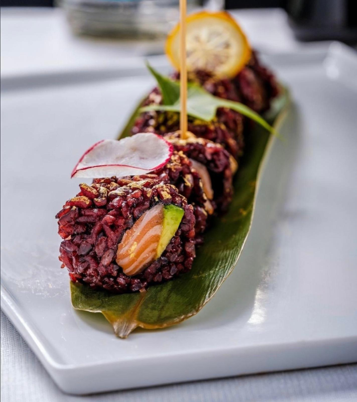Involtini di sushi su una foglia verde, guarnita con una fetta di ravanello e una guarnizione arancione, su un piatto rettangolare bianco.
