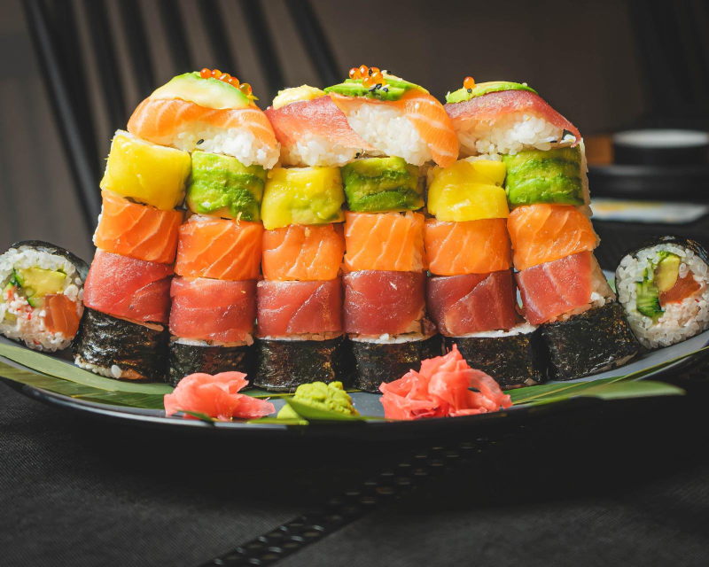 Torre di sushi con strati colorati: salmone, tonno, mango, avocado, su un piatto con zenzero e wasabi.