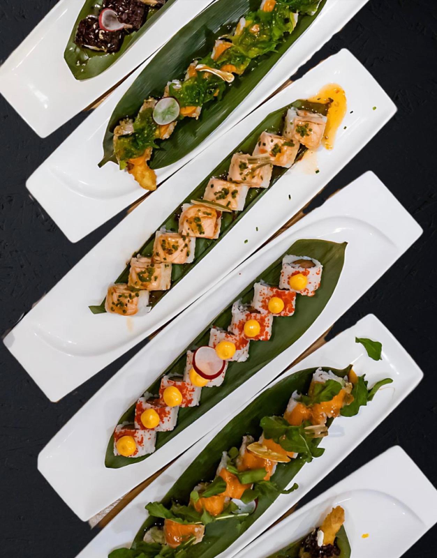 Involtini di sushi disposti su vassoi bianchi, sopra grandi foglie verdi su sfondo nero.