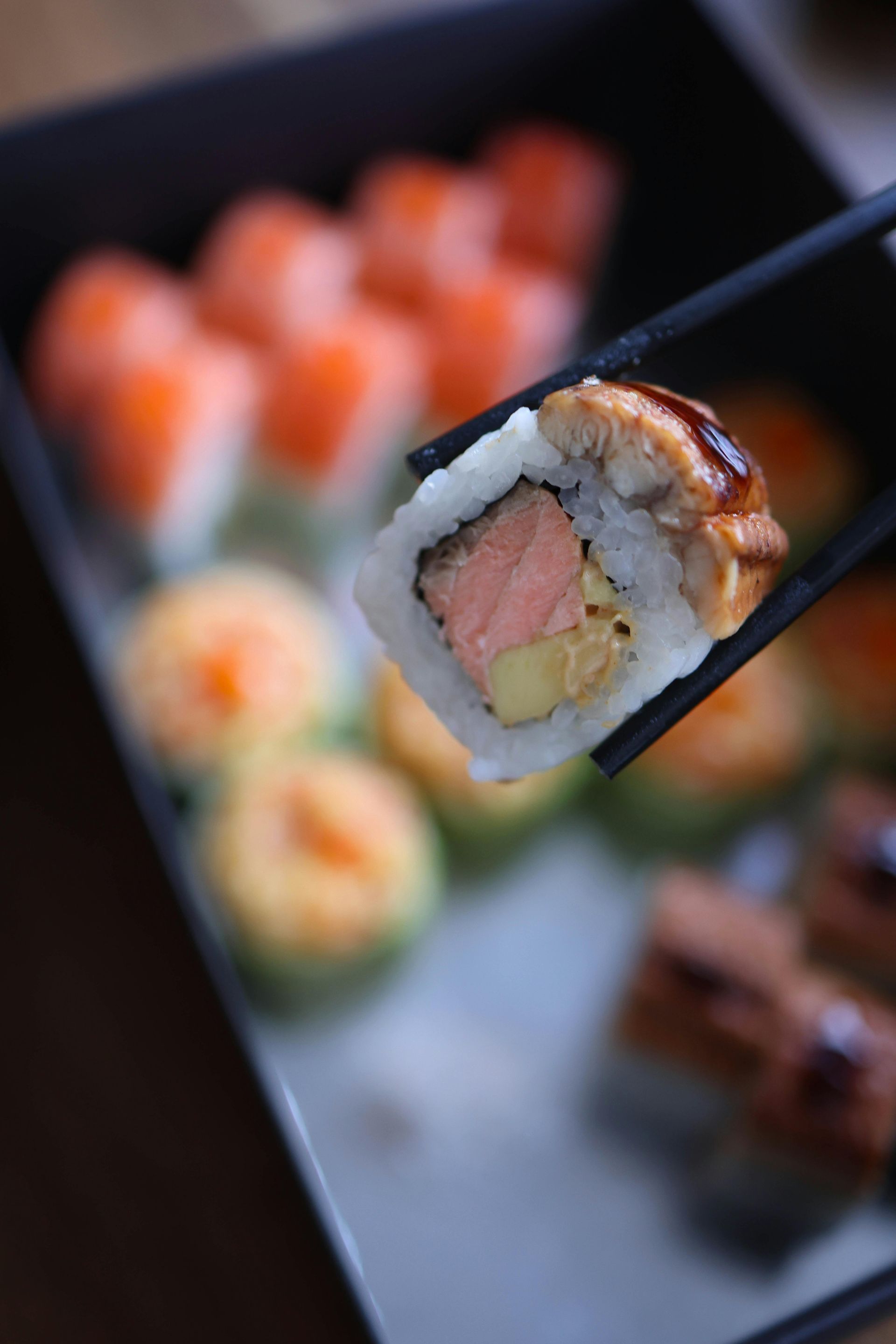 Rotolo di sushi tenuto con le bacchette, scatola di panini sullo sfondo.