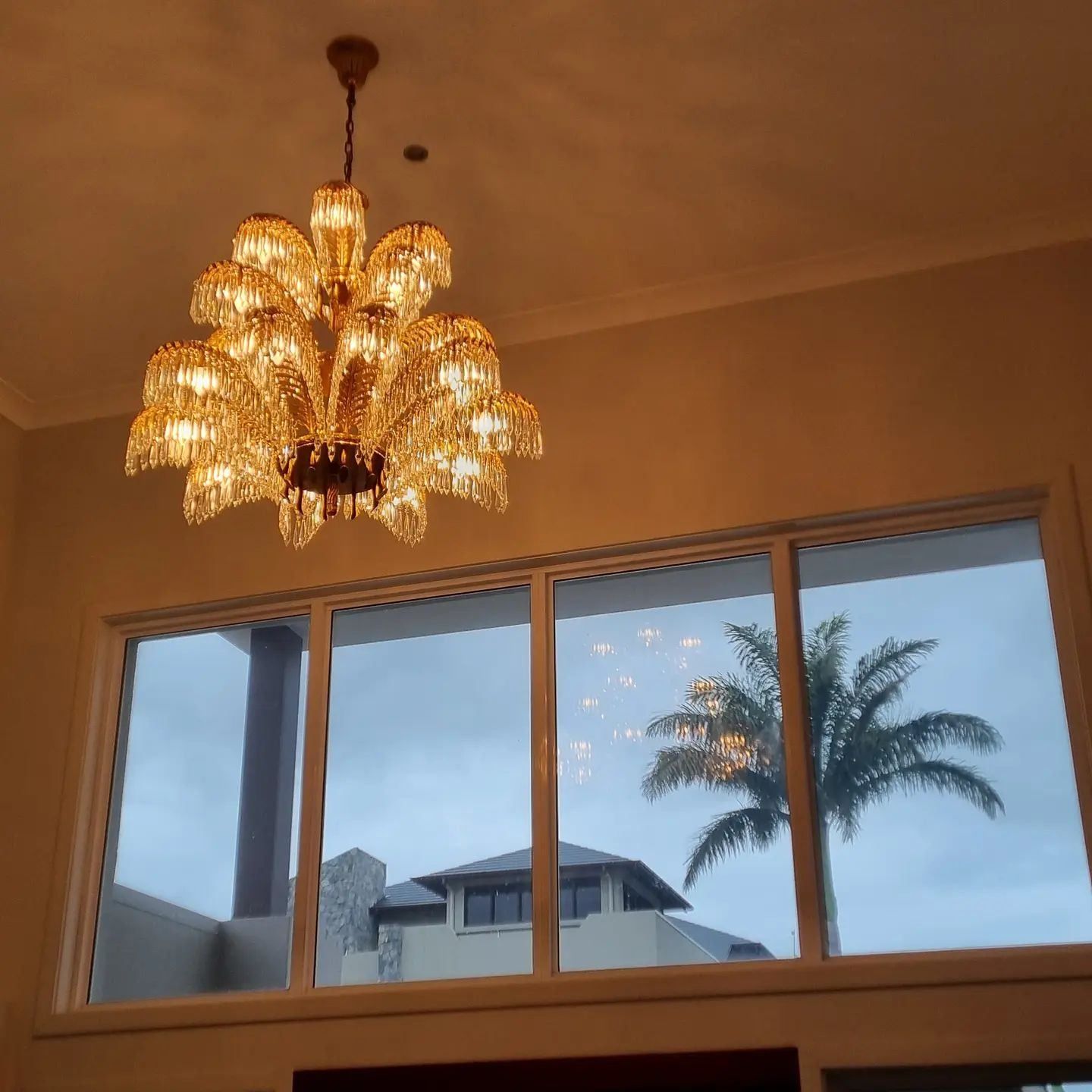 A Chandelier Hangs from The Ceiling Above a Window with Palm Trees in The Background — Goldspark GC Electrical in Burleigh Waters, QLD