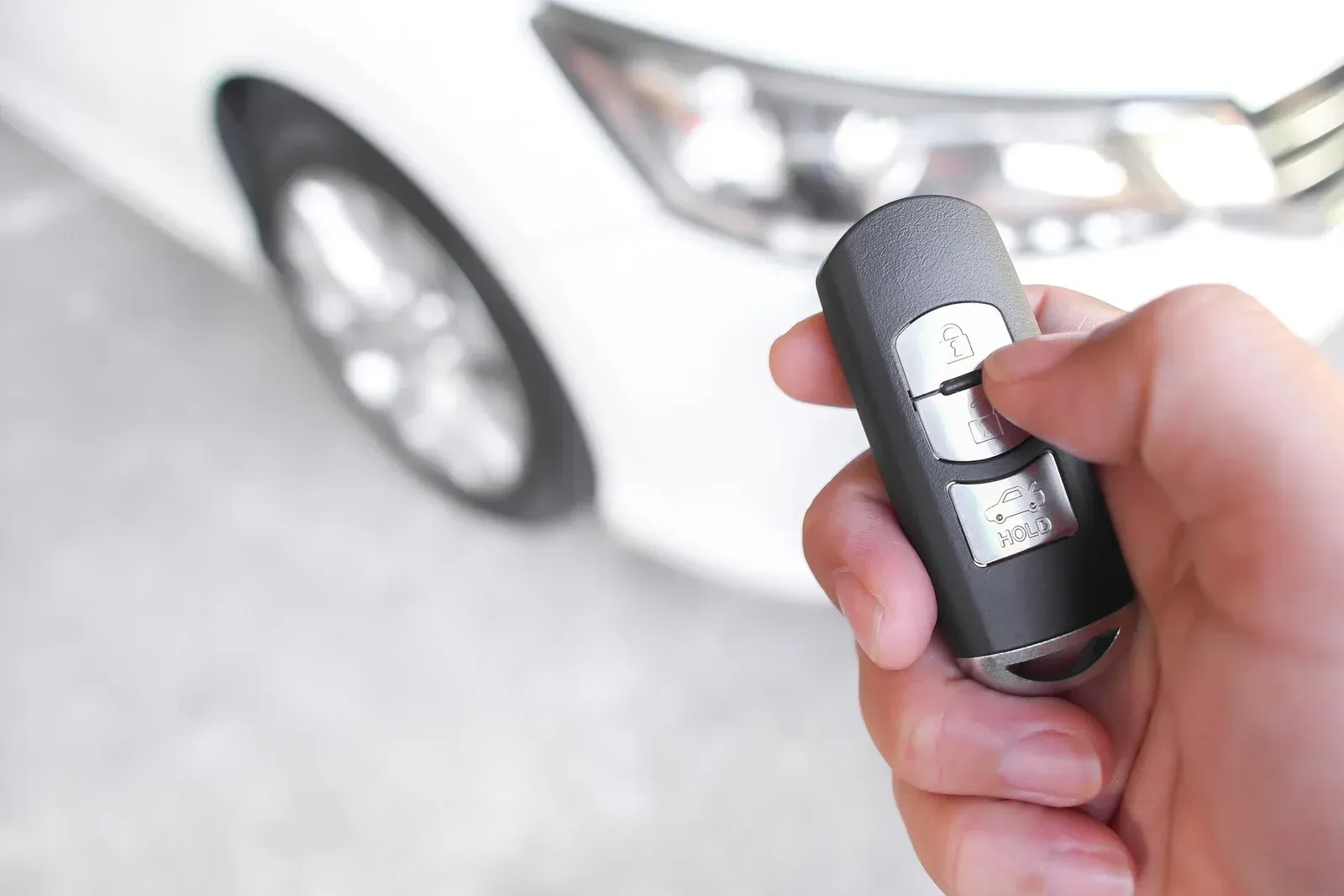 A hand holding a car key fob to unlock a white vehicle parked in the background.