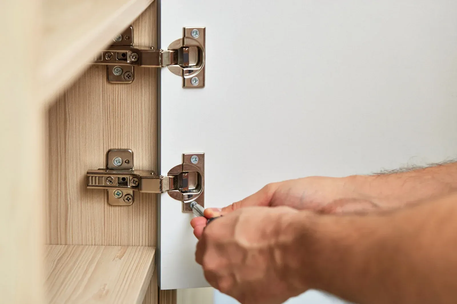 A person uses a screwdriver to adjust the hinge on a white cabinet door attached to a light-colored wood frame.