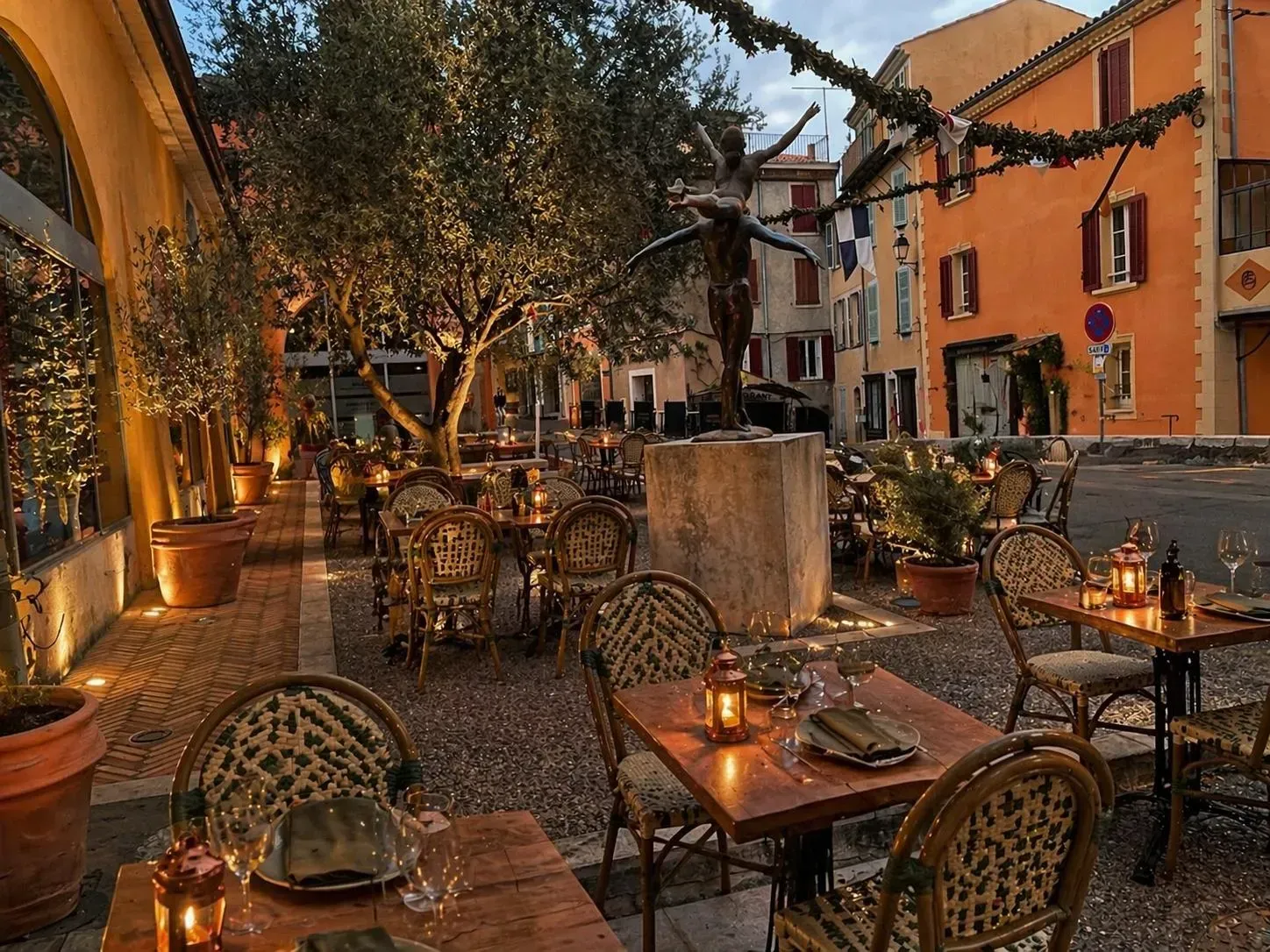 Terrasse de café en plein air accueillante au crépuscule, avec chaises en osier, tables, lumières chaudes et plantes en pot, dans une rue étroite.