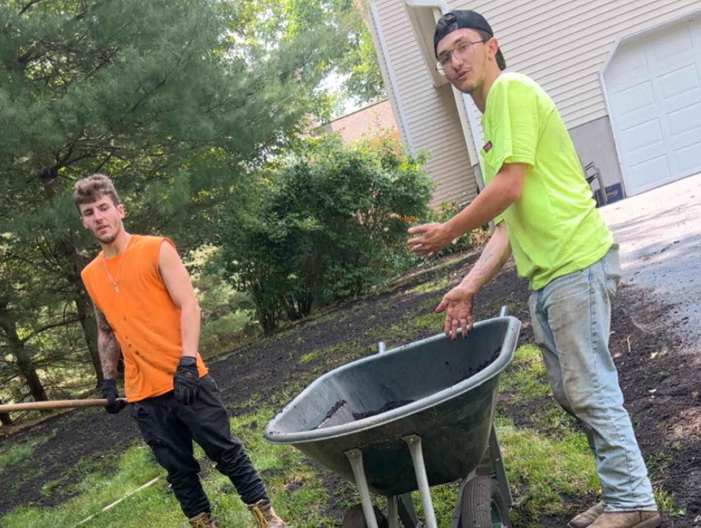 Two men are working in a yard with a wheelbarrow and shovel.