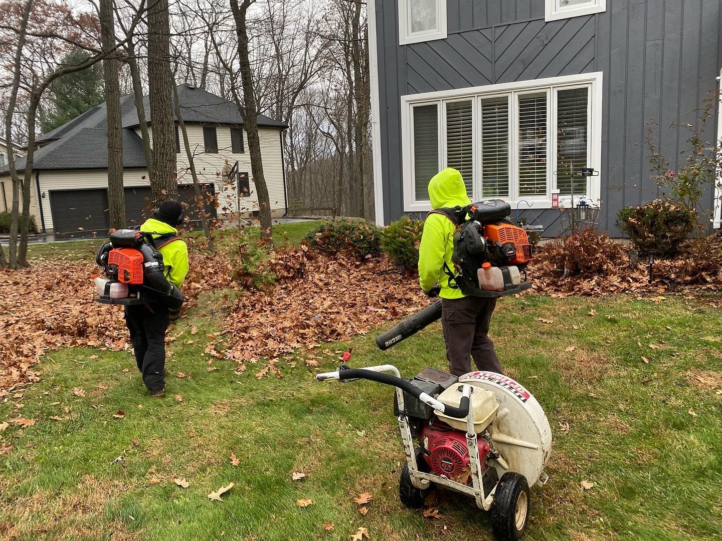 Two people are blowing leaves in front of a house.
