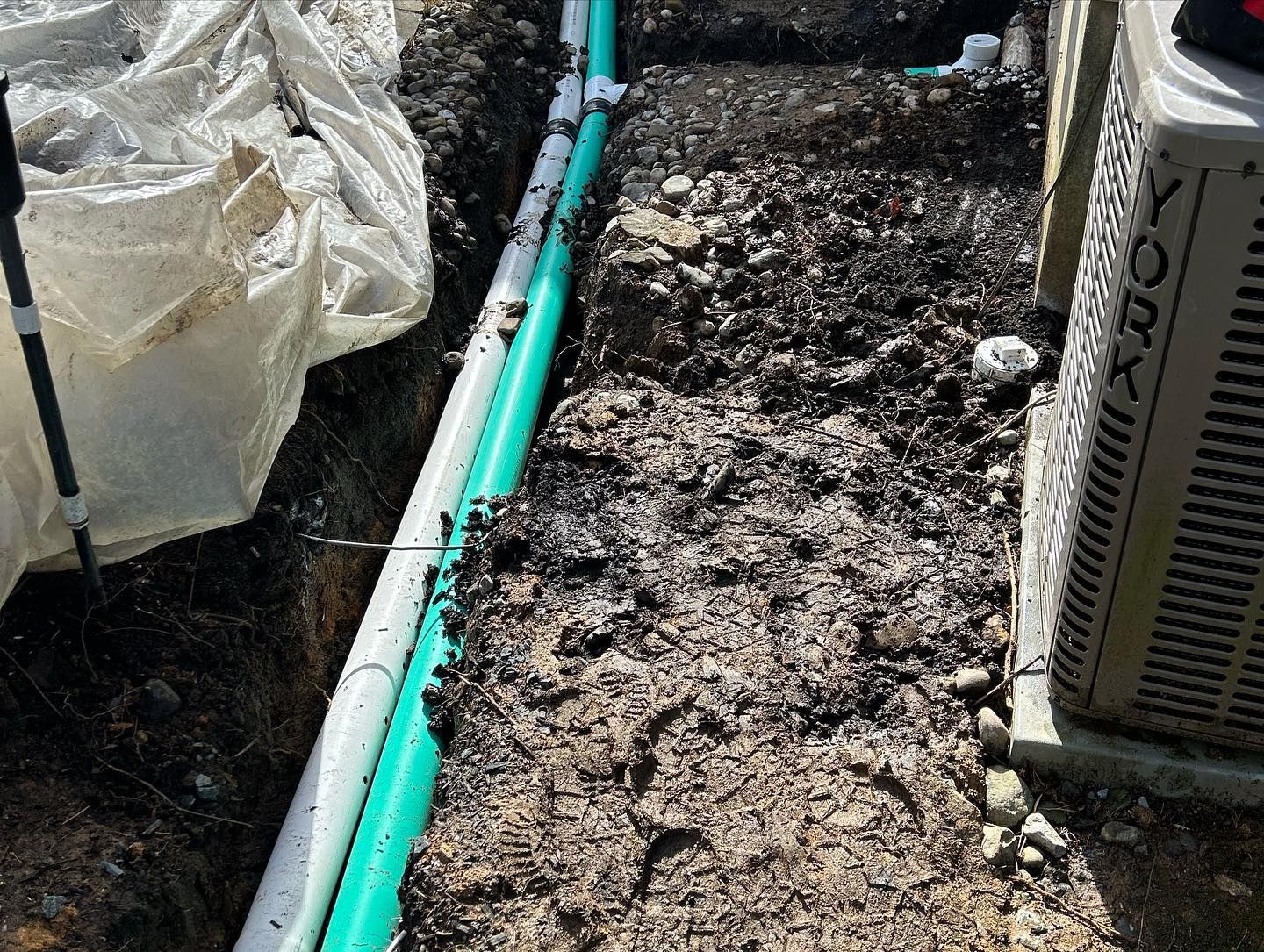 A york air conditioner is sitting in the dirt next to a green pipe.