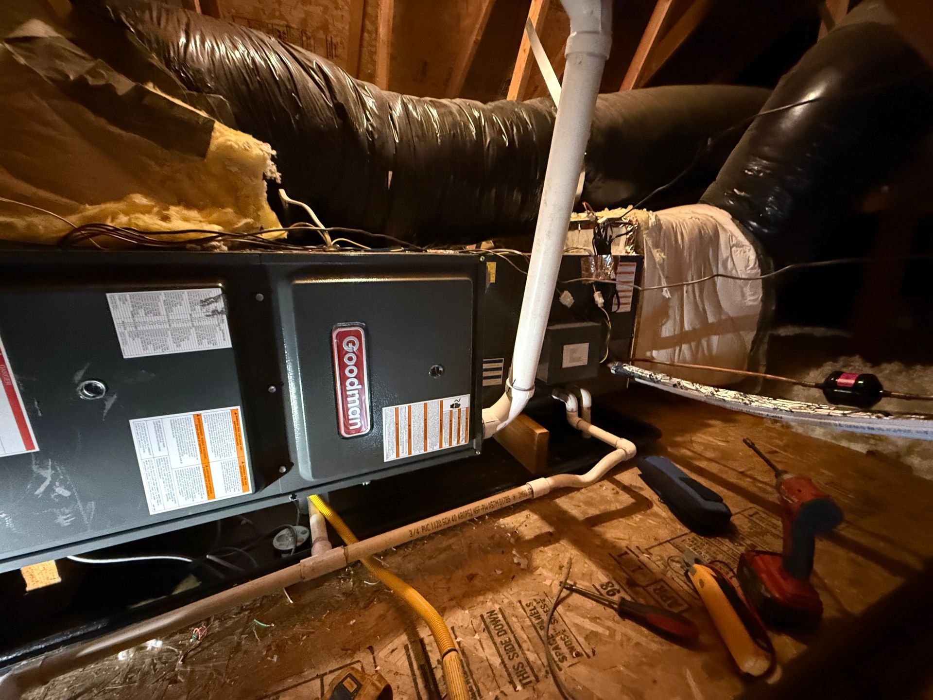 HVAC unit in an attic. Gray furnace with ductwork and plumbing. Tools on wooden floor.