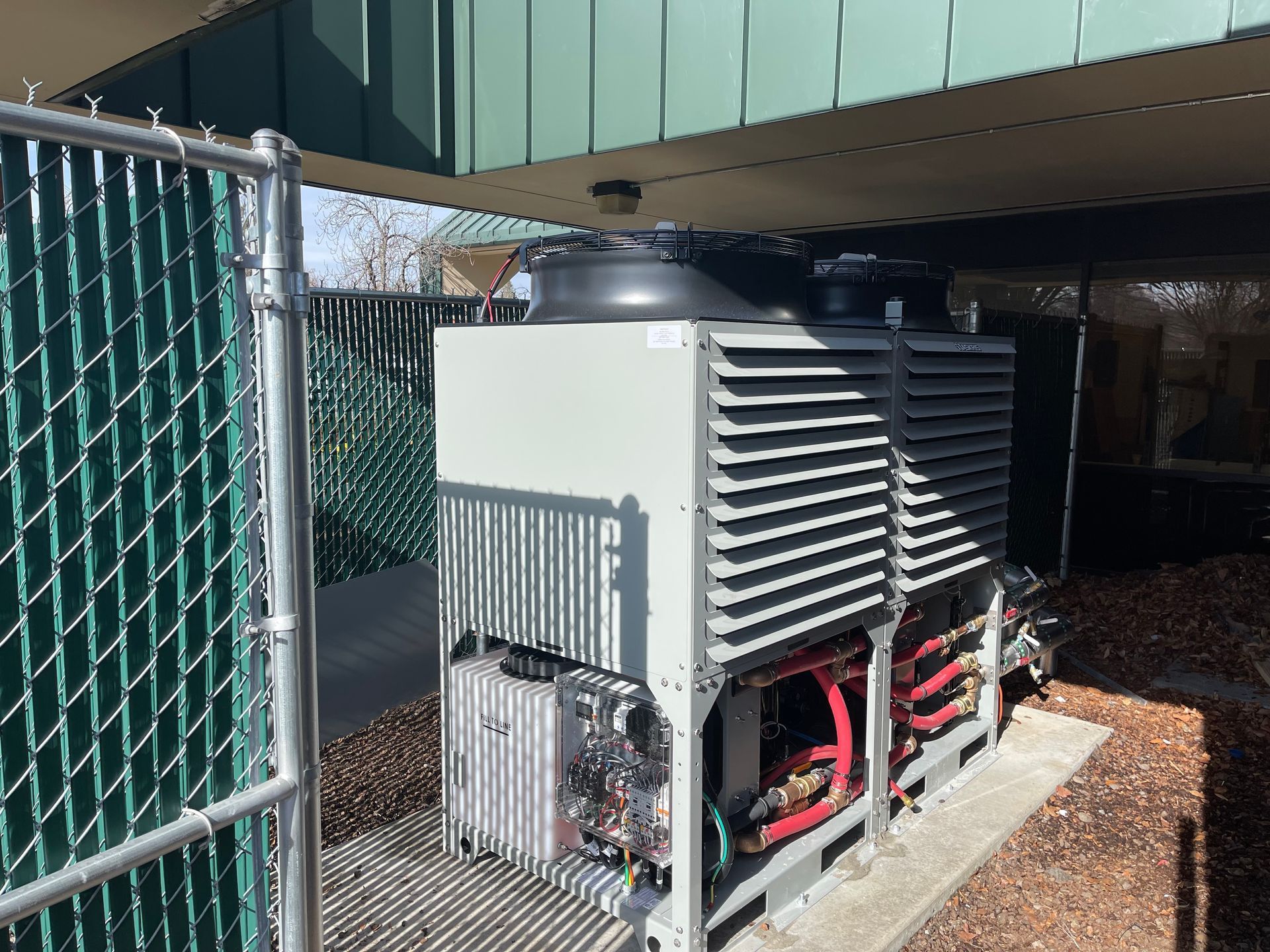 Two large industrial air conditioning units behind a chain-link fence, under a building overhang.