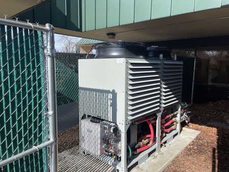 Two industrial HVAC units behind a chain-link fence, under a building overhang.