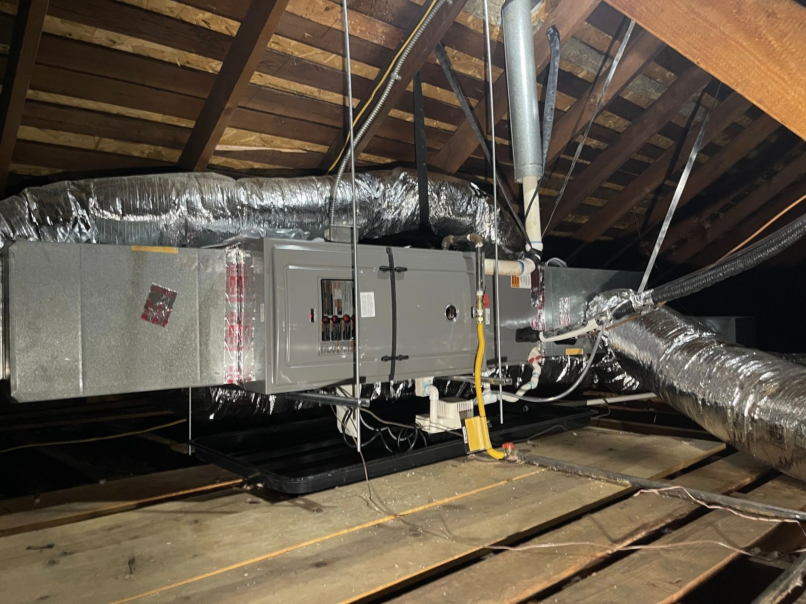 HVAC unit installed in an attic; metallic components, insulated ducts, electrical wiring, wooden beams.