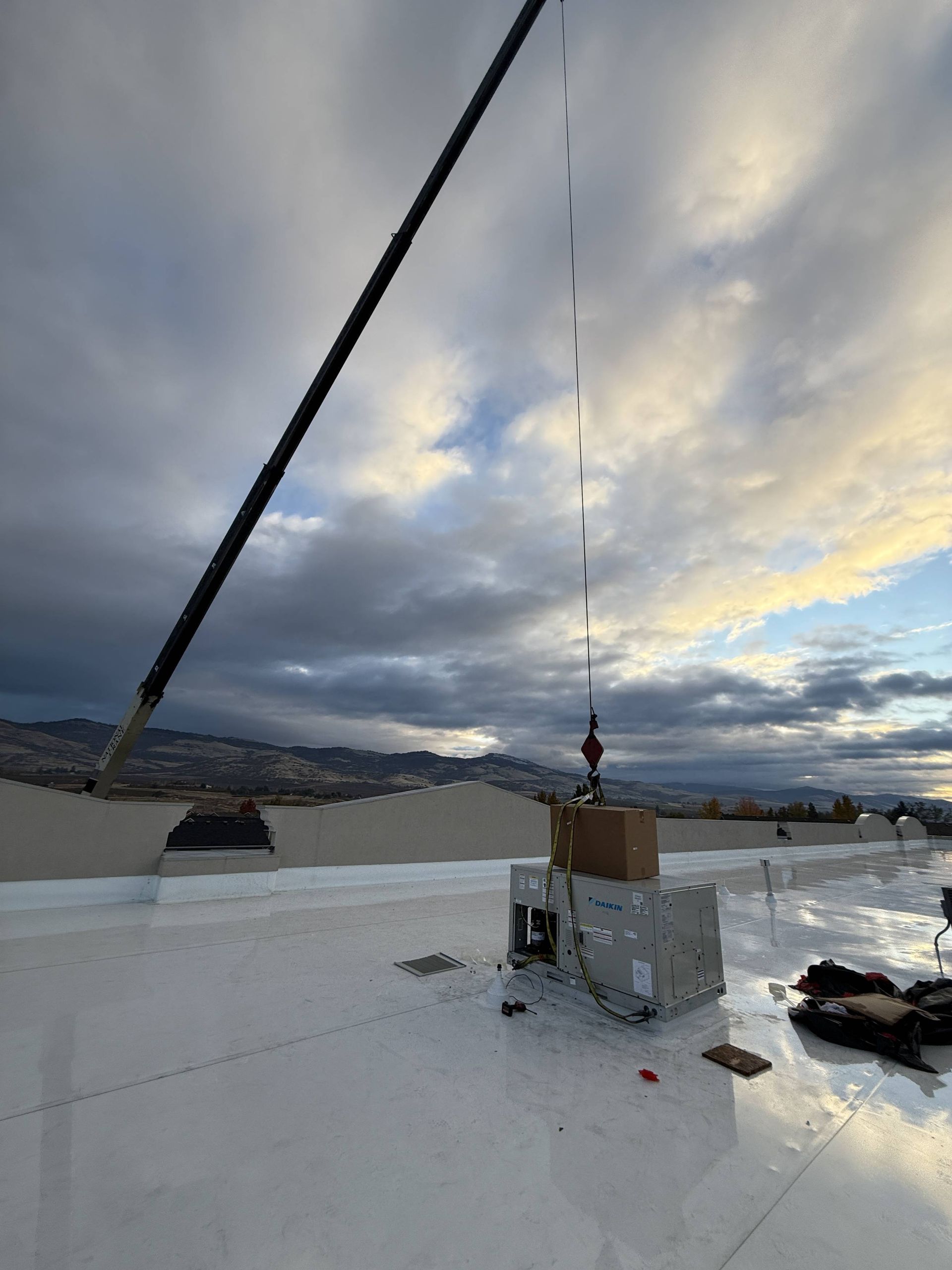 Crane lifting a box onto a white rooftop with mountains in the background under a cloudy sky.