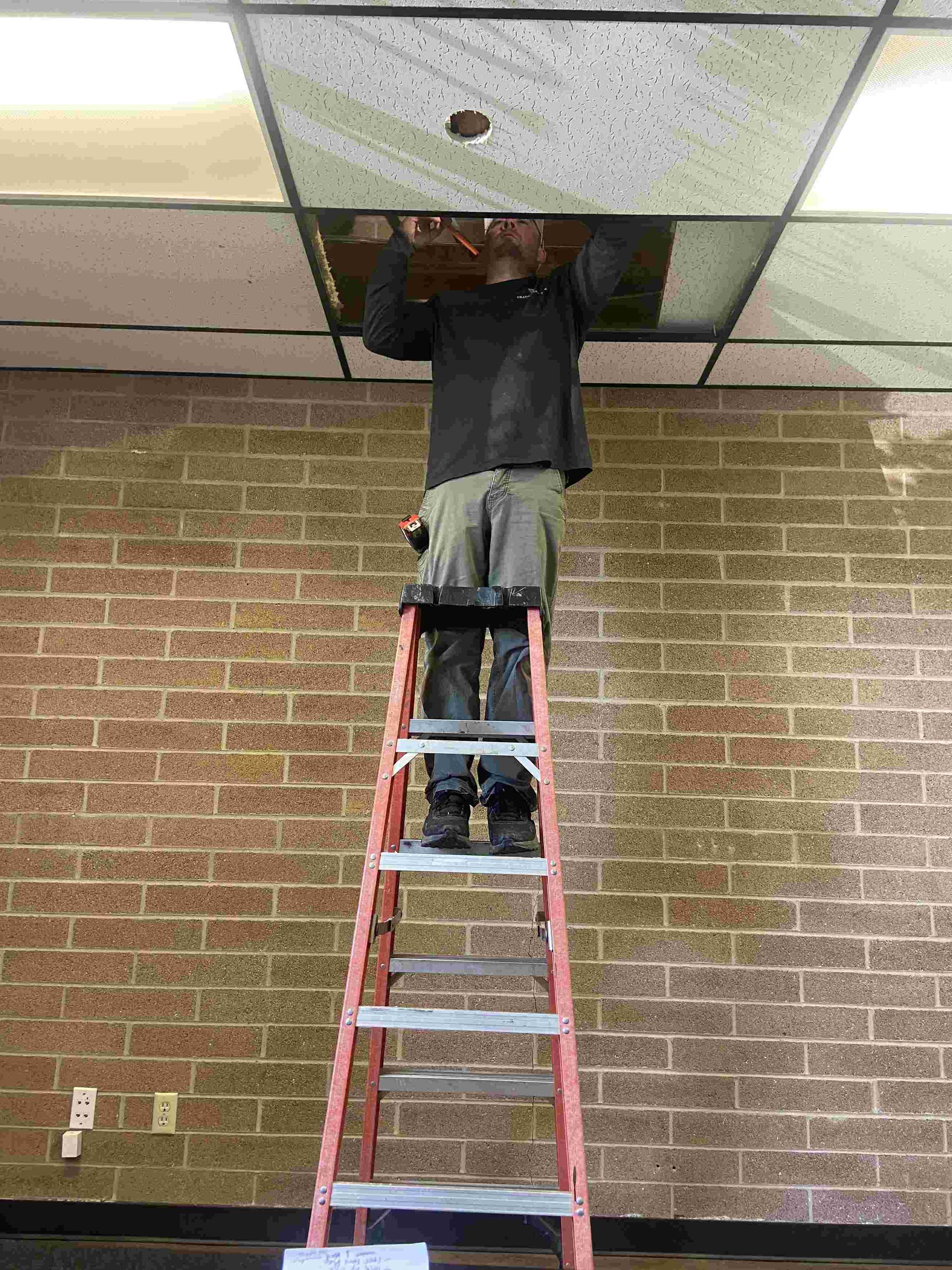 Person on ladder working on a ceiling tile. Red ladder, brick wall background.