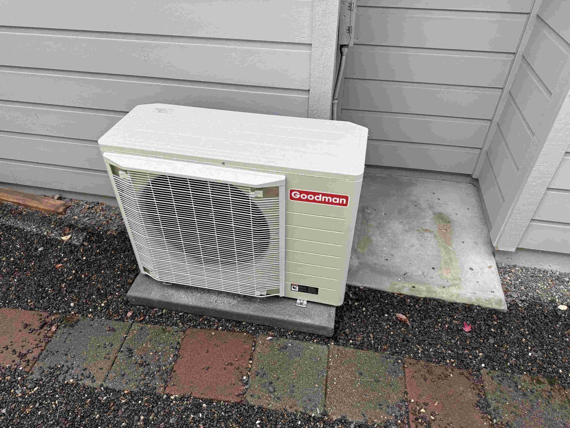 Outdoor Goodman air conditioning unit on a concrete pad next to a house with gray siding.