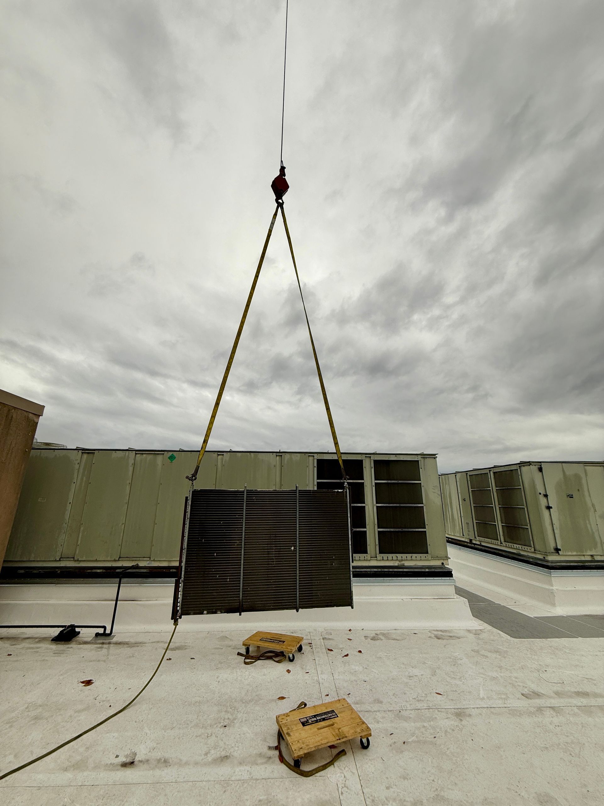 Crane lifting an HVAC unit on a building rooftop under a cloudy sky.