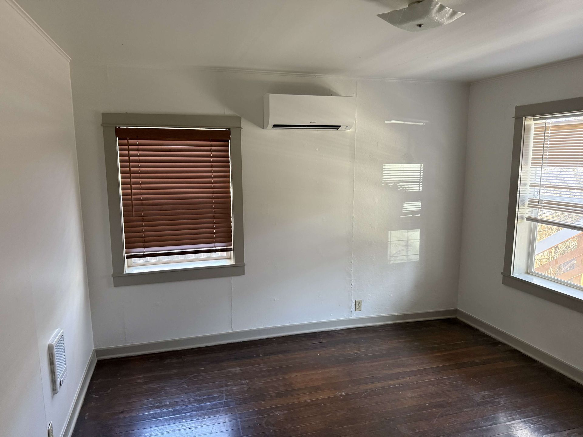 Empty room with dark wood floors, white walls, two windows, and an air conditioning unit.