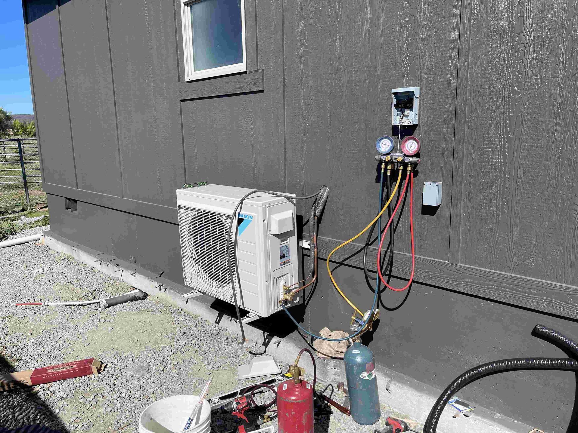 An HVAC technician servicing an outdoor air conditioning unit connected to gauges and tanks; exterior of a building.