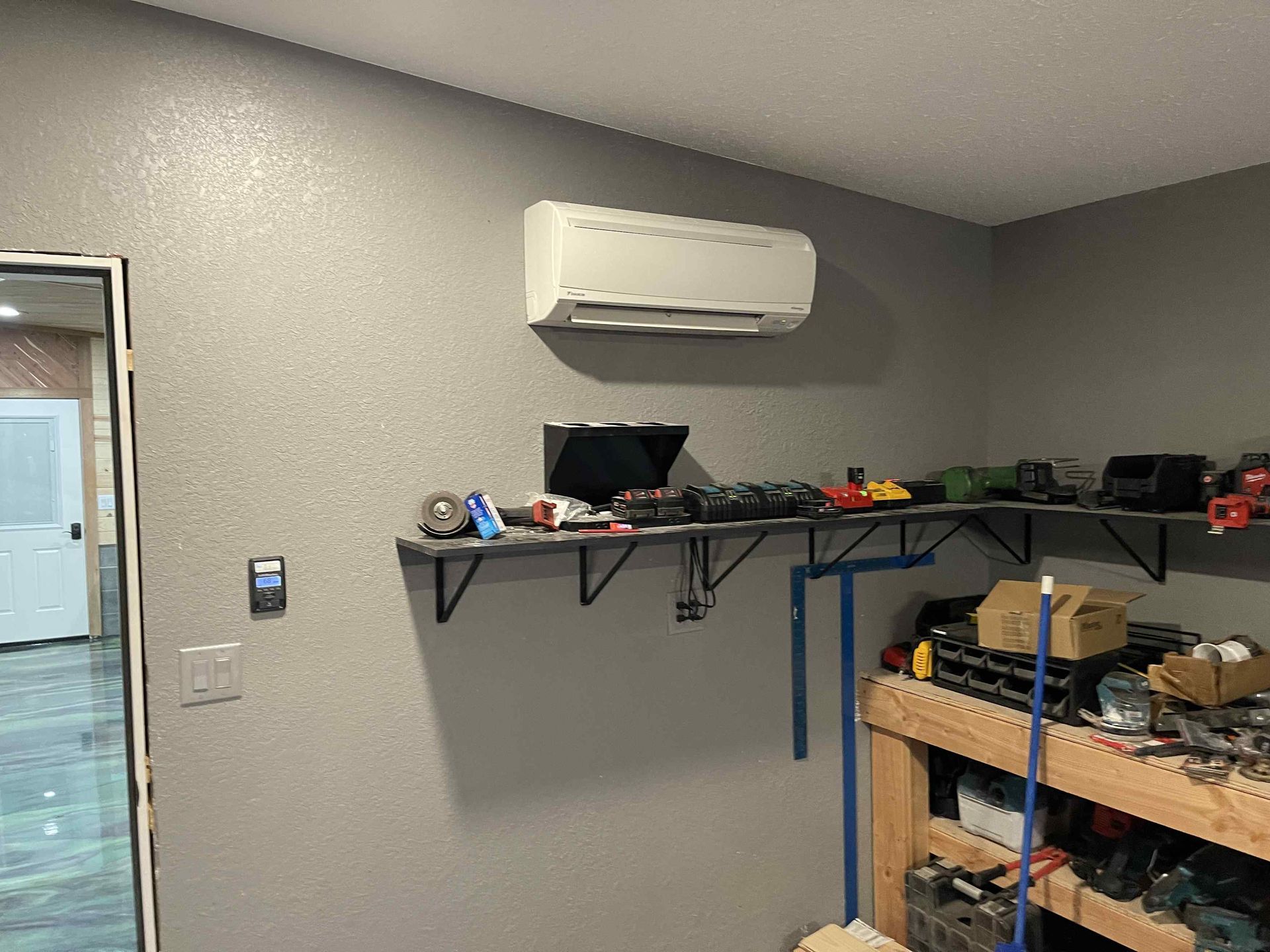 Wall-mounted air conditioner above a shelf with tools and equipment in a garage. Gray walls, white ceiling.