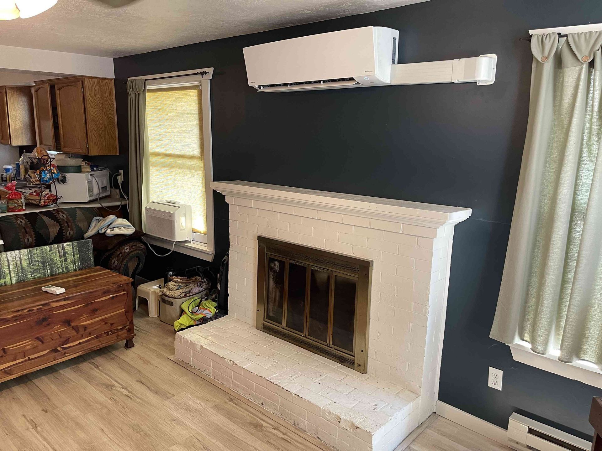 White fireplace, dark blue wall with air conditioner, window, wooden cabinets, and a wooden chest in a room.
