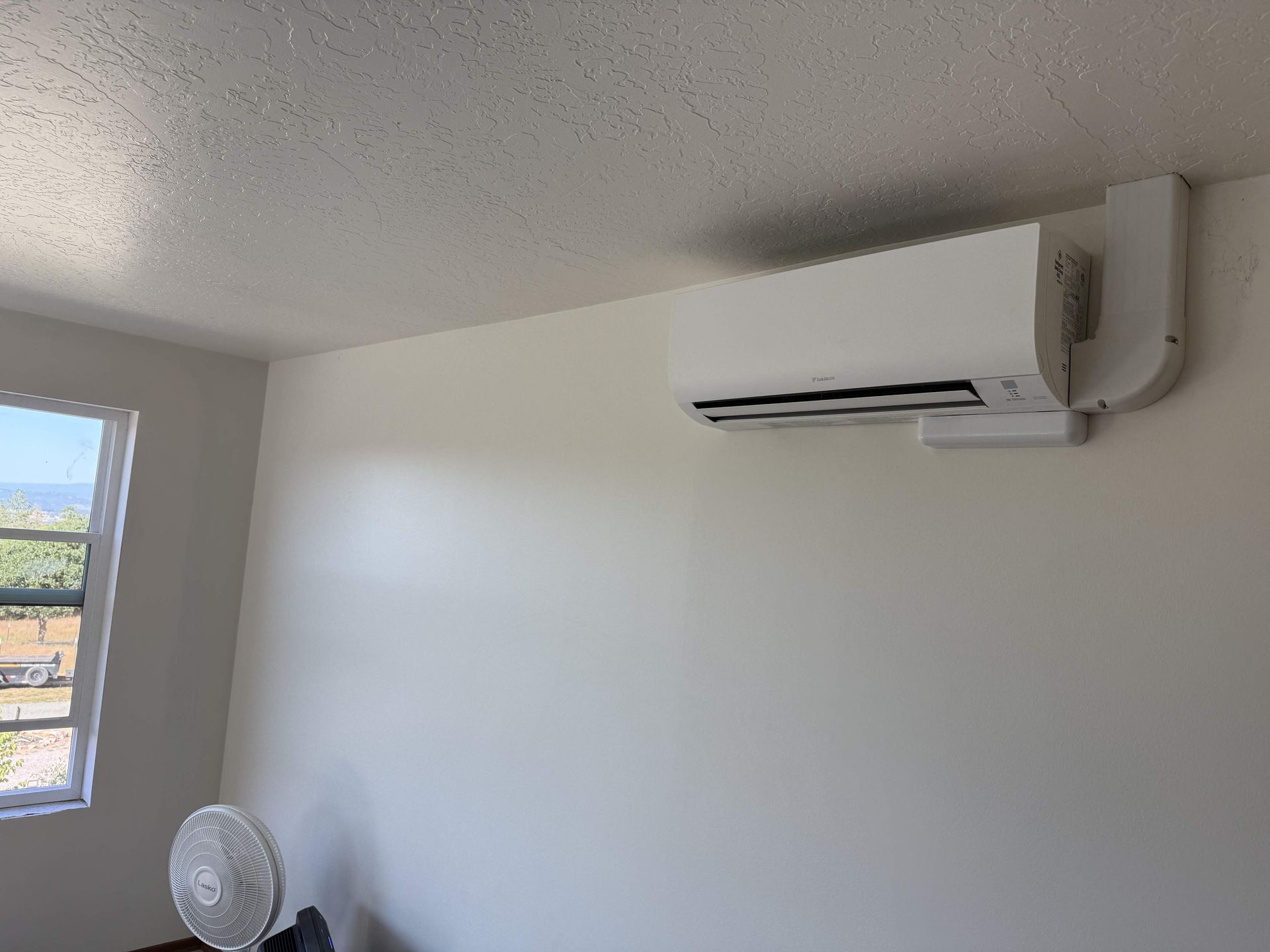 White air conditioner mounted on a white wall in a room, with a window and a fan visible.