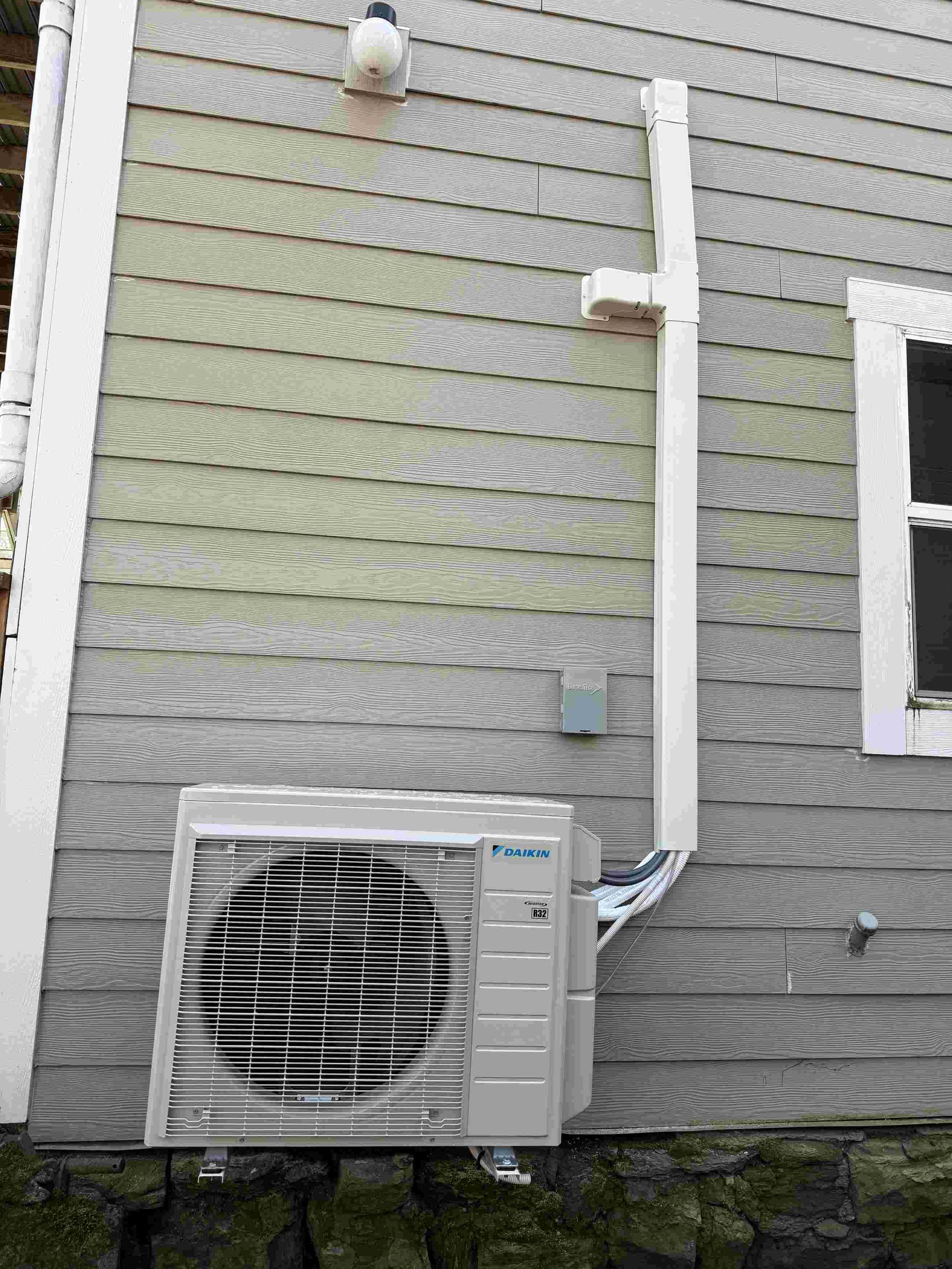 Outdoor air conditioning unit mounted on a gray-sided house with a white conduit.