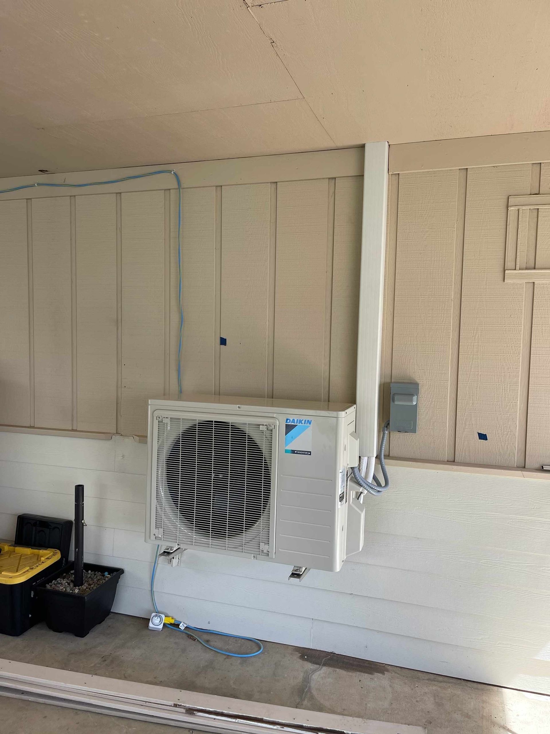 White mini-split AC unit mounted on a light beige siding wall. White conduit runs up to a gray electrical box.