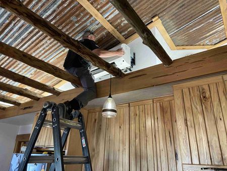 Man on ladder installing light fixture near wood beams and corrugated metal ceiling.