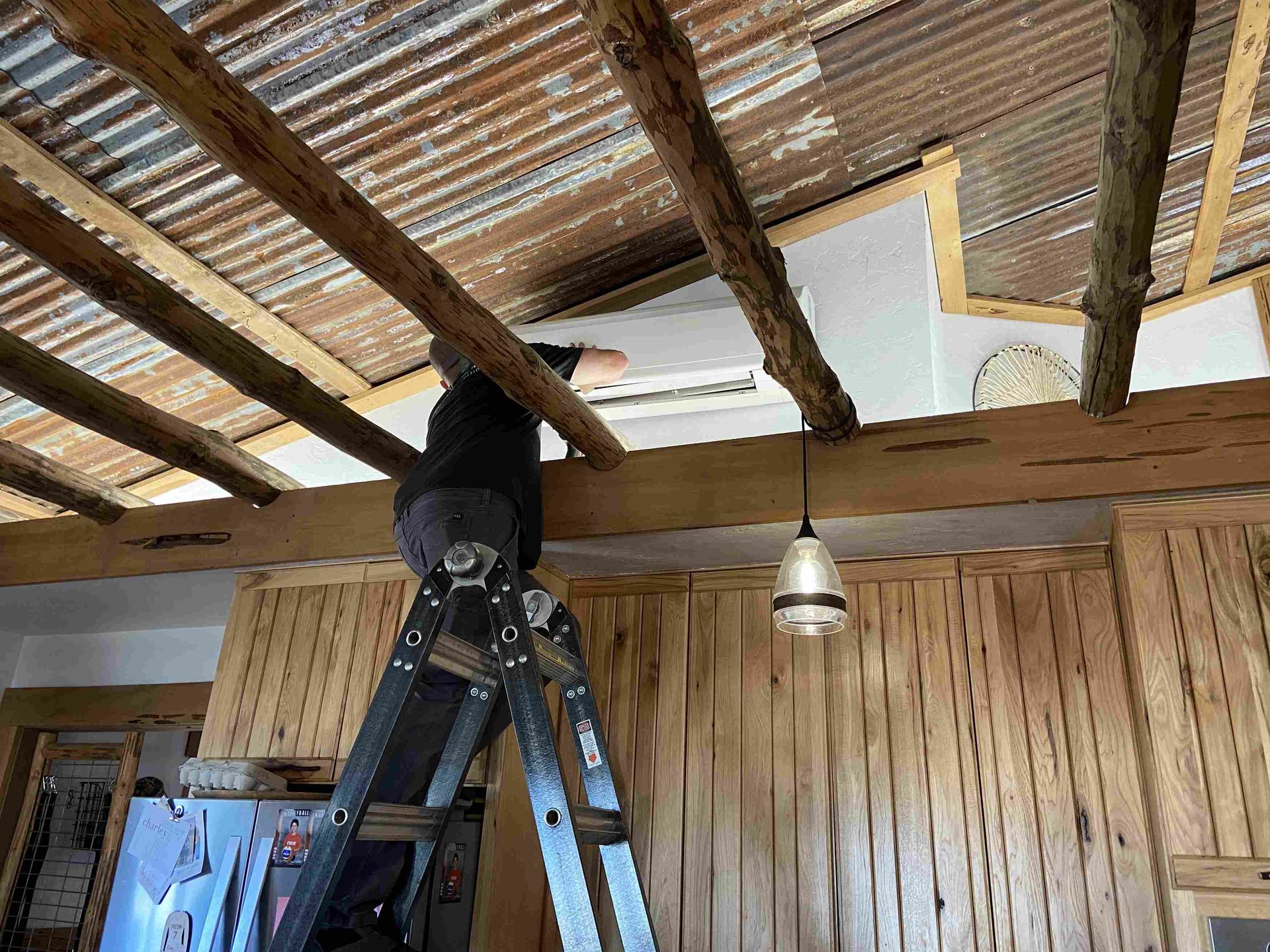 Person on ladder reaching toward a ceiling vent in a rustic room with exposed beams.