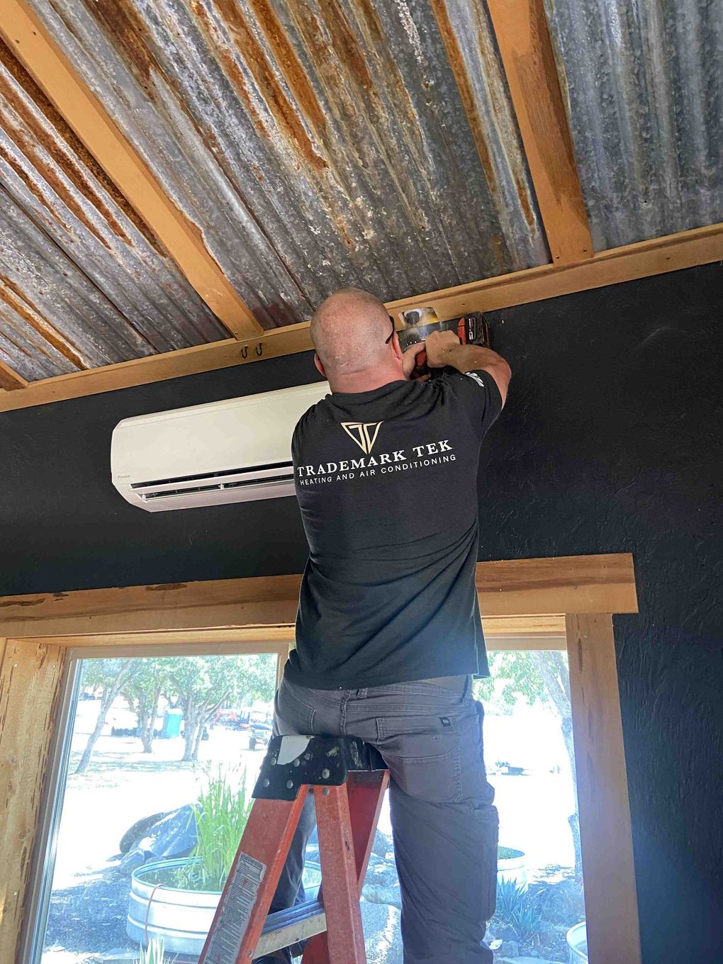 Man on a ladder installing an AC unit on a black wall, under a corrugated metal ceiling.