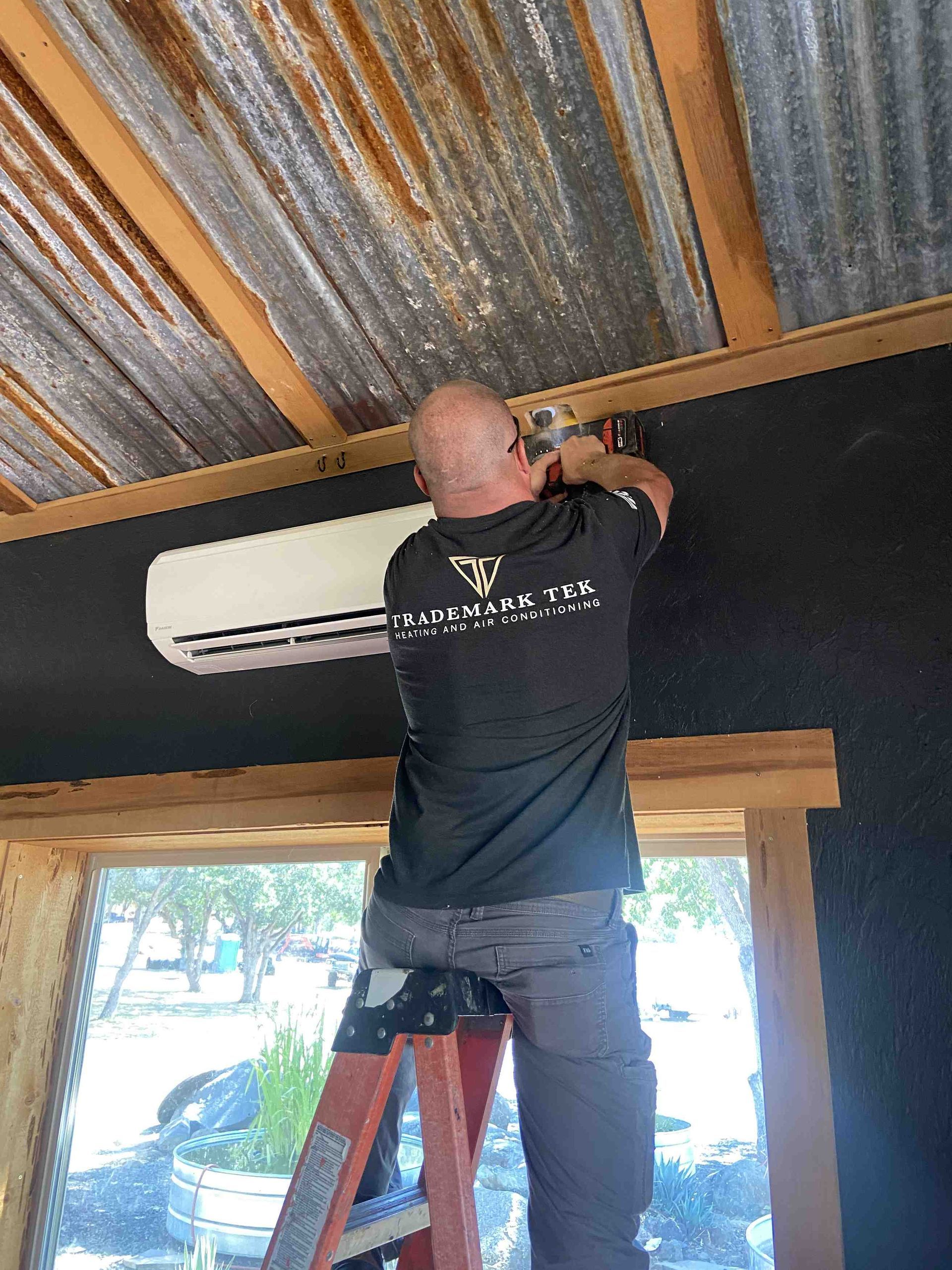 Man on ladder installing an AC unit inside a building with a corrugated metal ceiling.