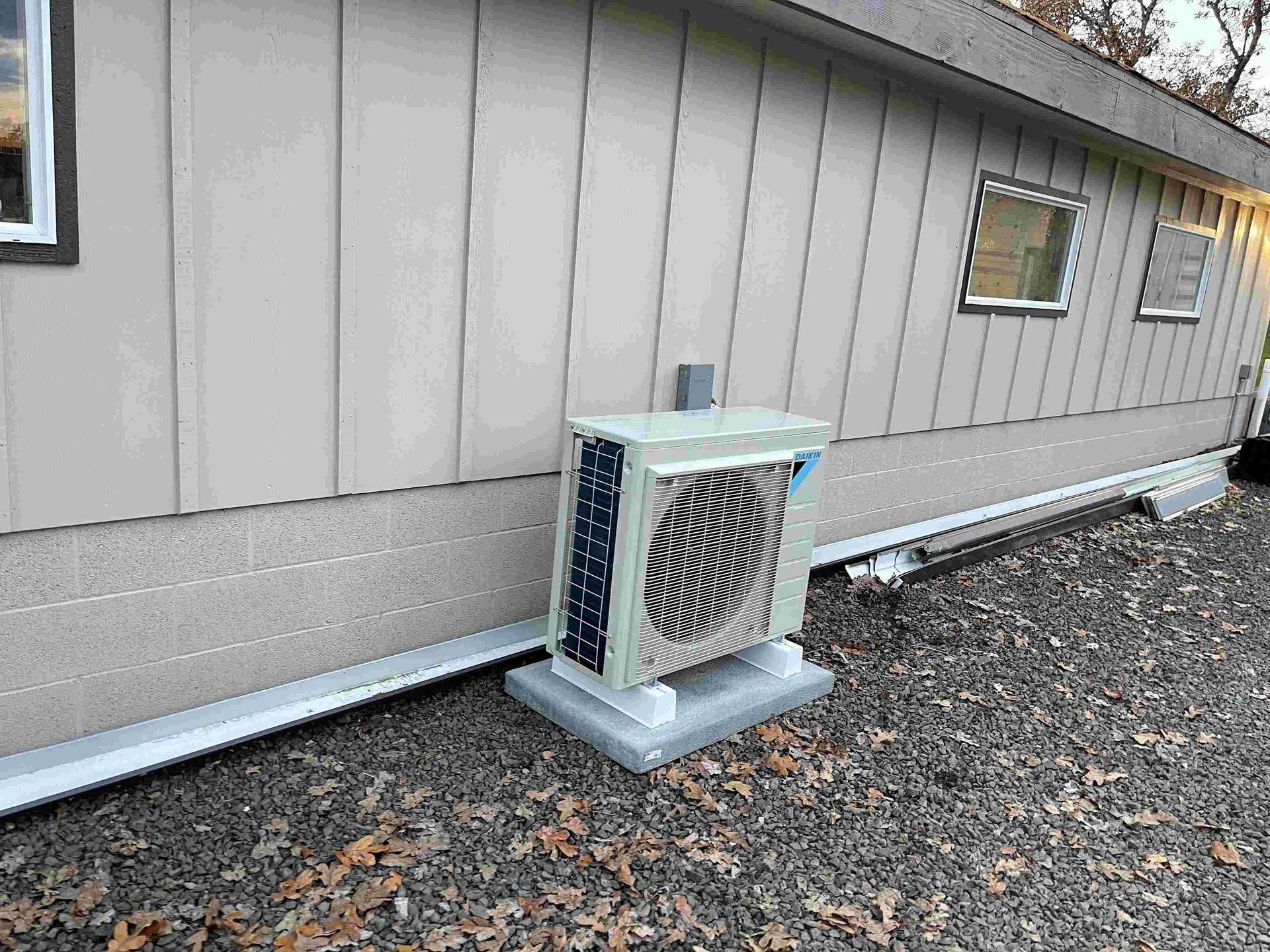 Air conditioning unit outside a beige building, sitting on a concrete pad, on a gravel surface.
