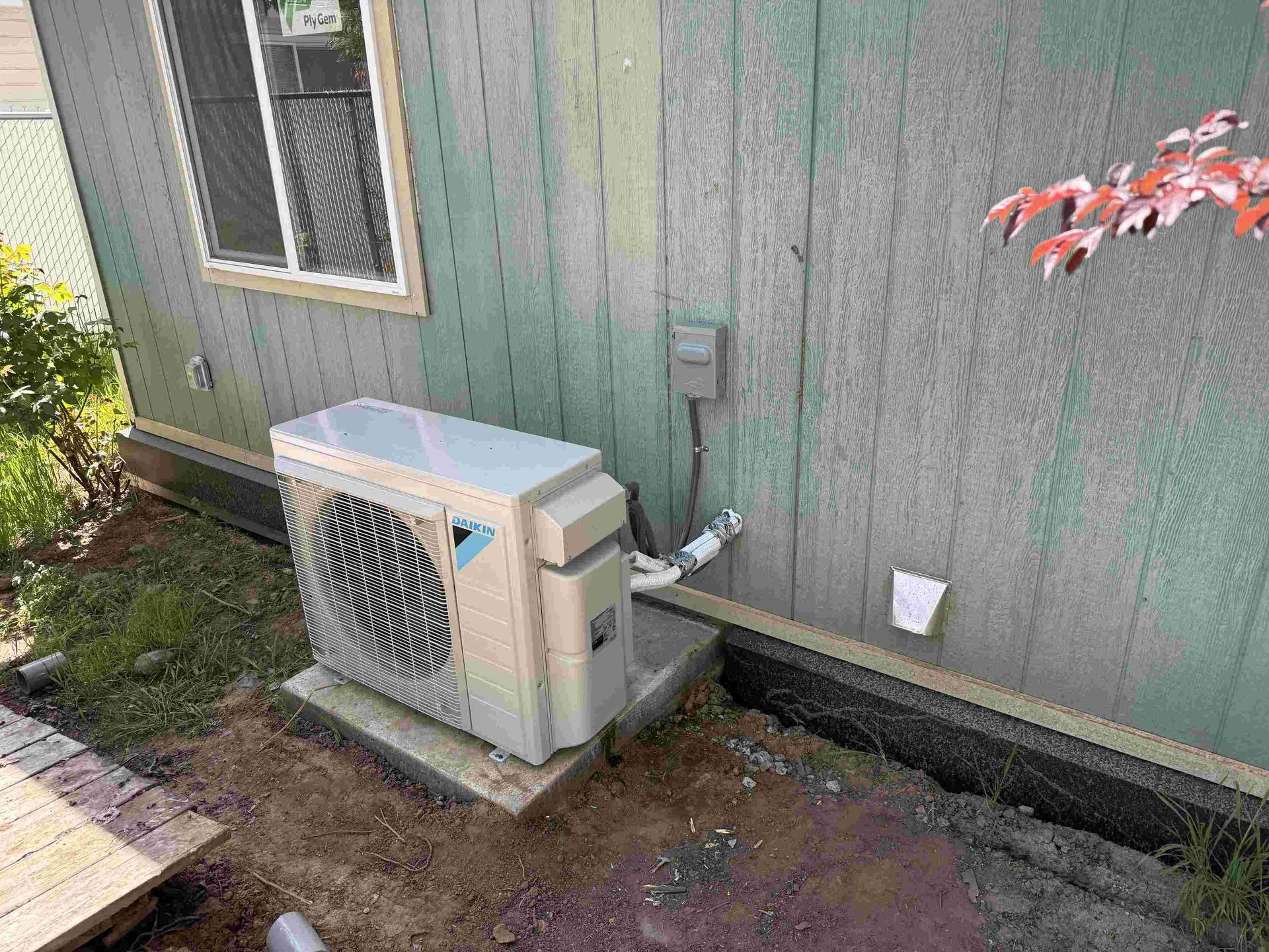 An air conditioning unit sits on a concrete pad next to a weathered building with a window.
