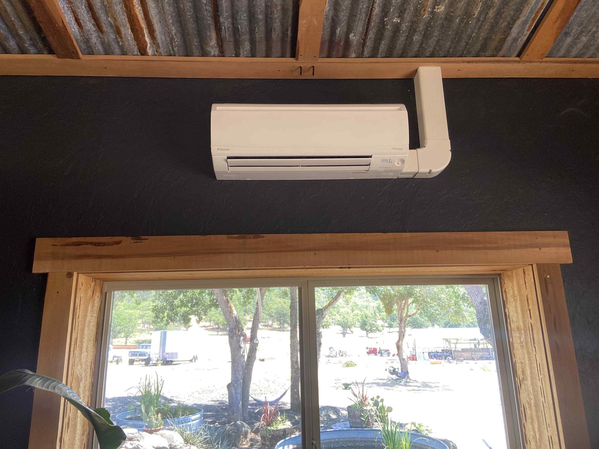 Air conditioner mounted on a dark wall above a window with a view of trees and sky.