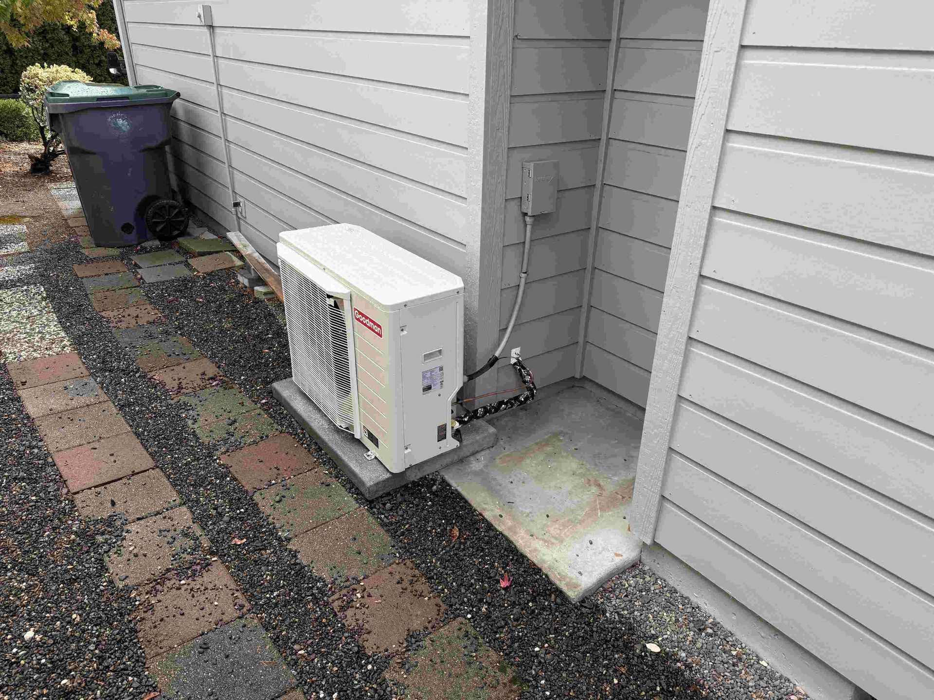 Outdoor air conditioning unit on a concrete pad next to a house with a trash can in view.