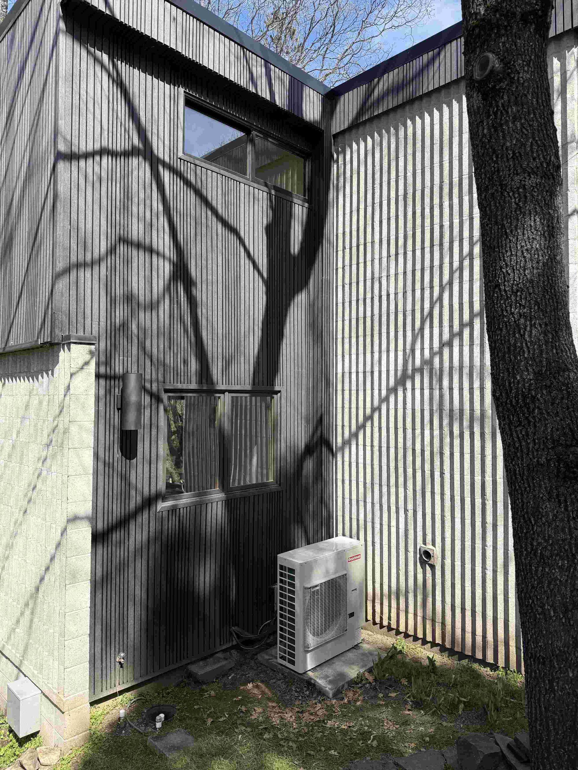 Exterior view of a building with vertical siding, window, and air conditioning unit. Tree shadows on wall.