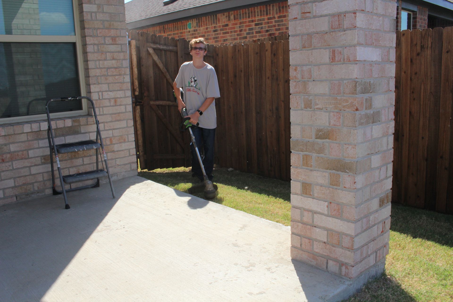 A young man is using a weed eater in a backyard next to a wooden fence.
