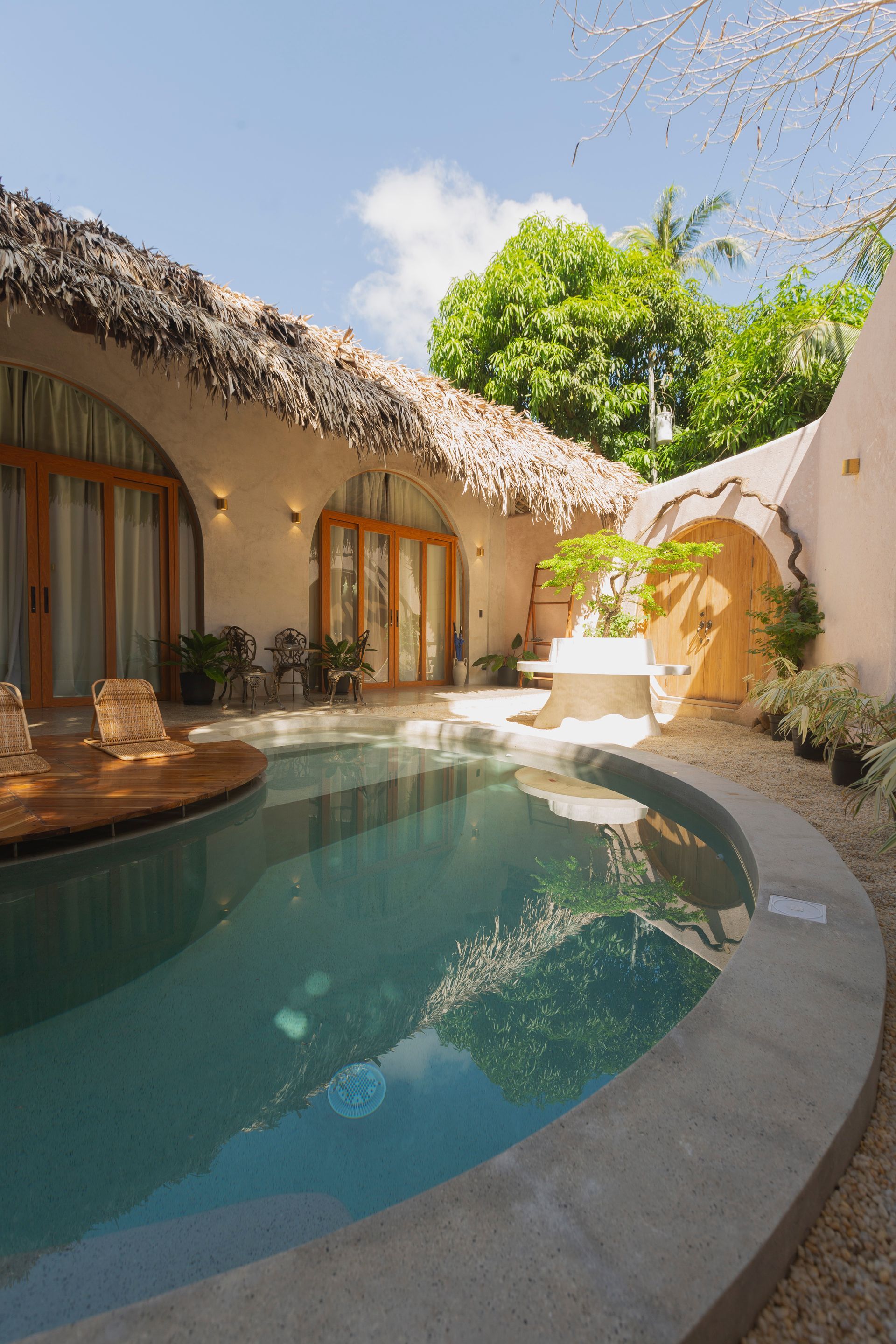 Pool in a tropical setting, next to a building with a thatched roof. Blue water, beige walls, sunny.