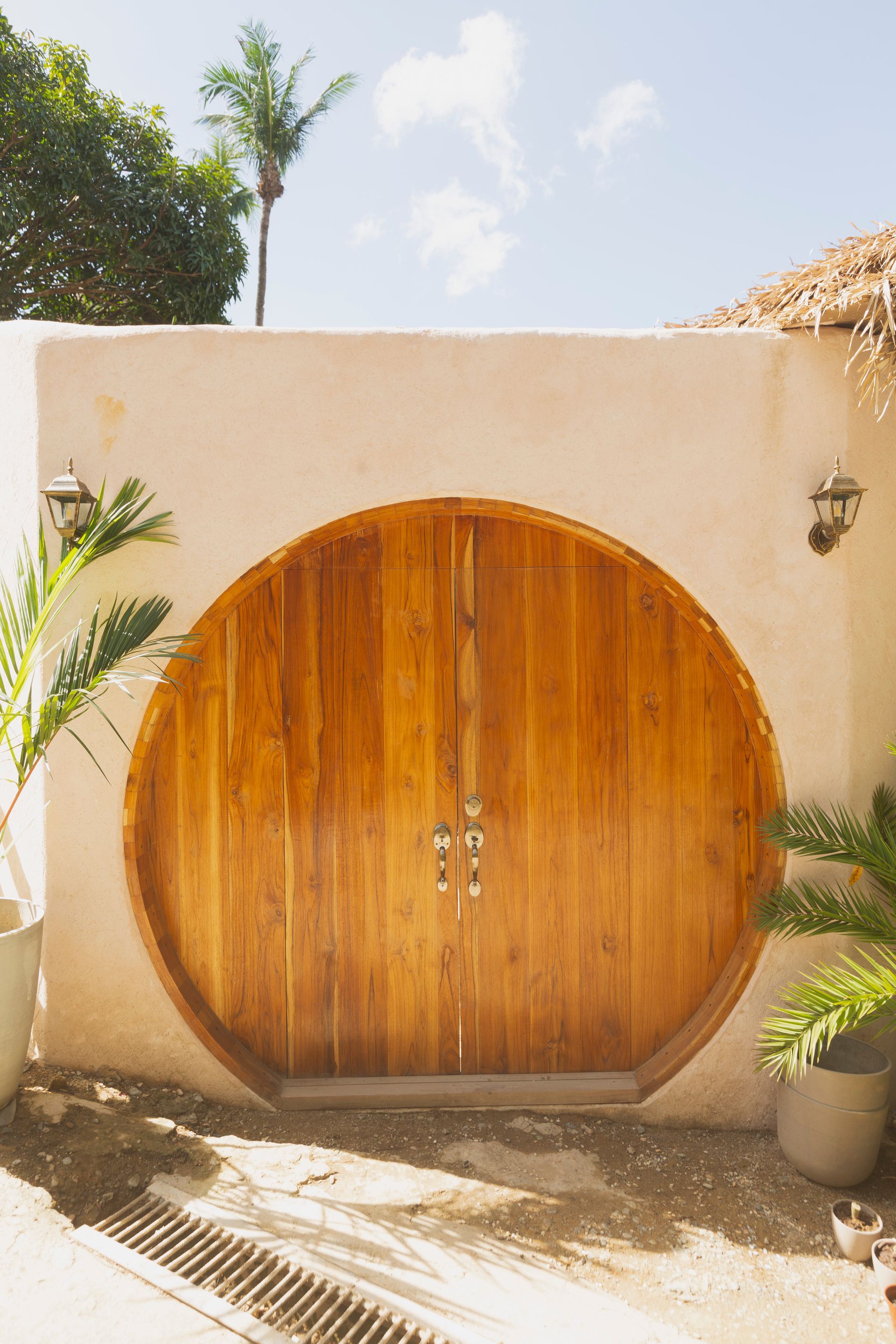 Wooden, circular double doors set in a cream-colored wall, flanked by greenery under a bright sky.