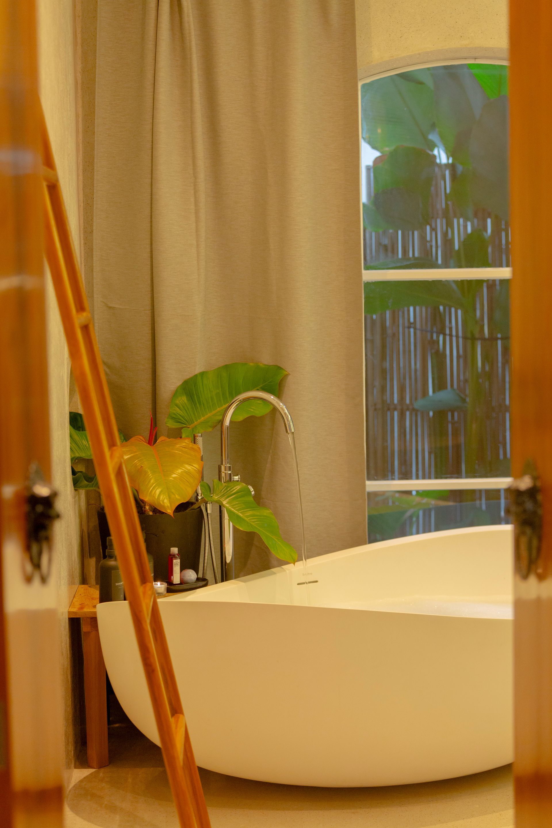 Oval bathtub by a window with greenery and tan curtains.