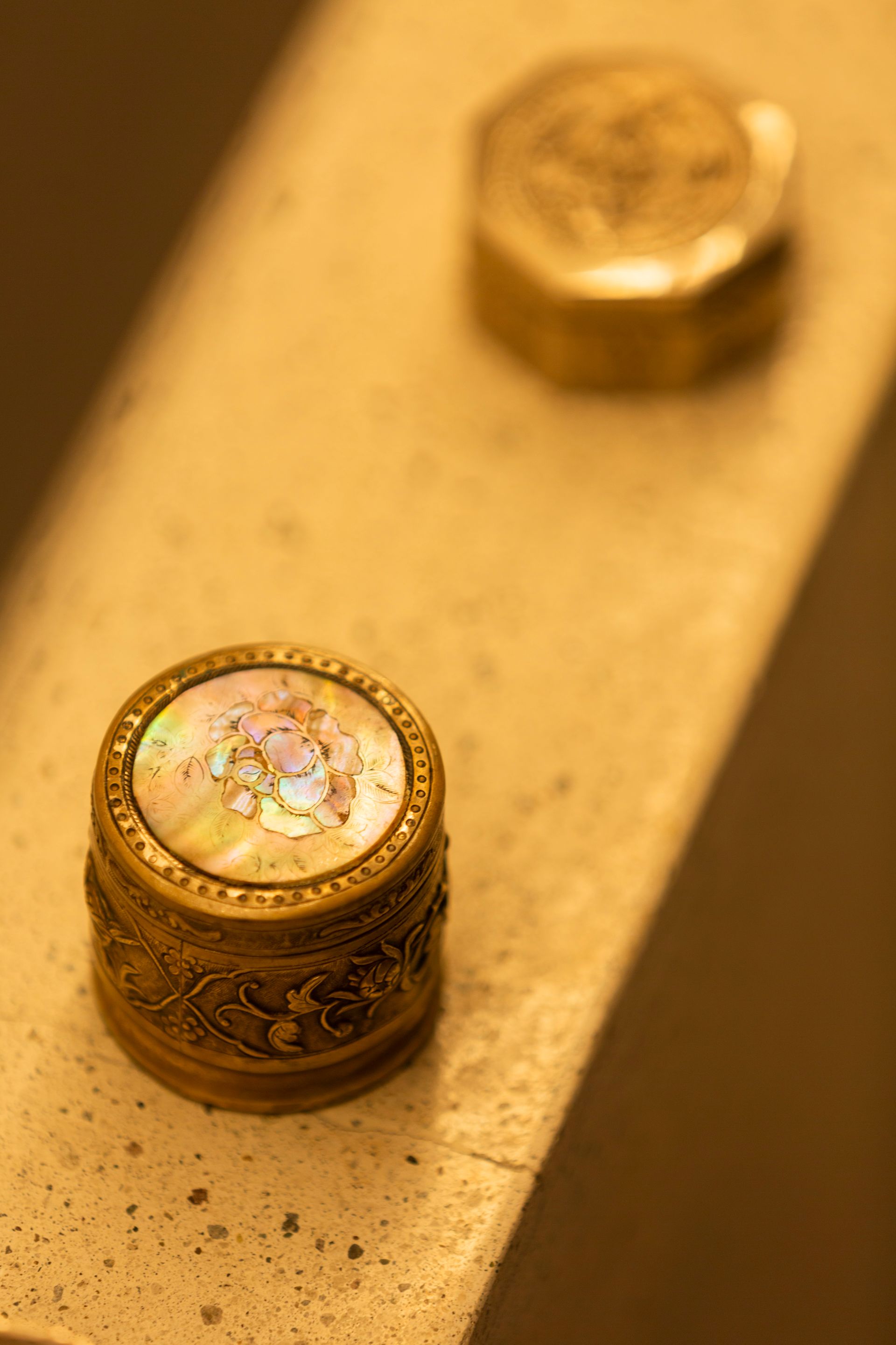 Two ornate, round, golden boxes on a light-colored surface; one with a pearl-like design on top.