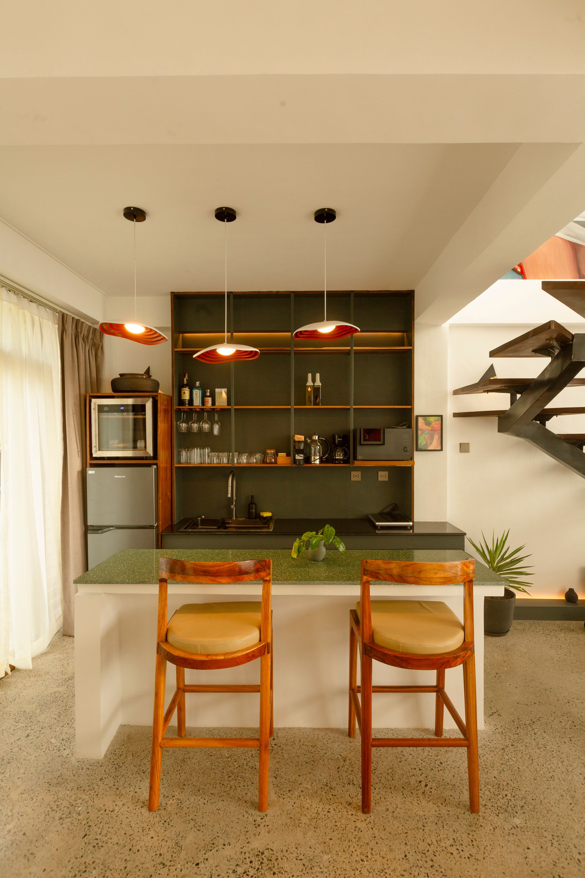 Interior with a bar, two stools, and a dark green shelving unit. White walls, speckled floor, and three pendant lights.