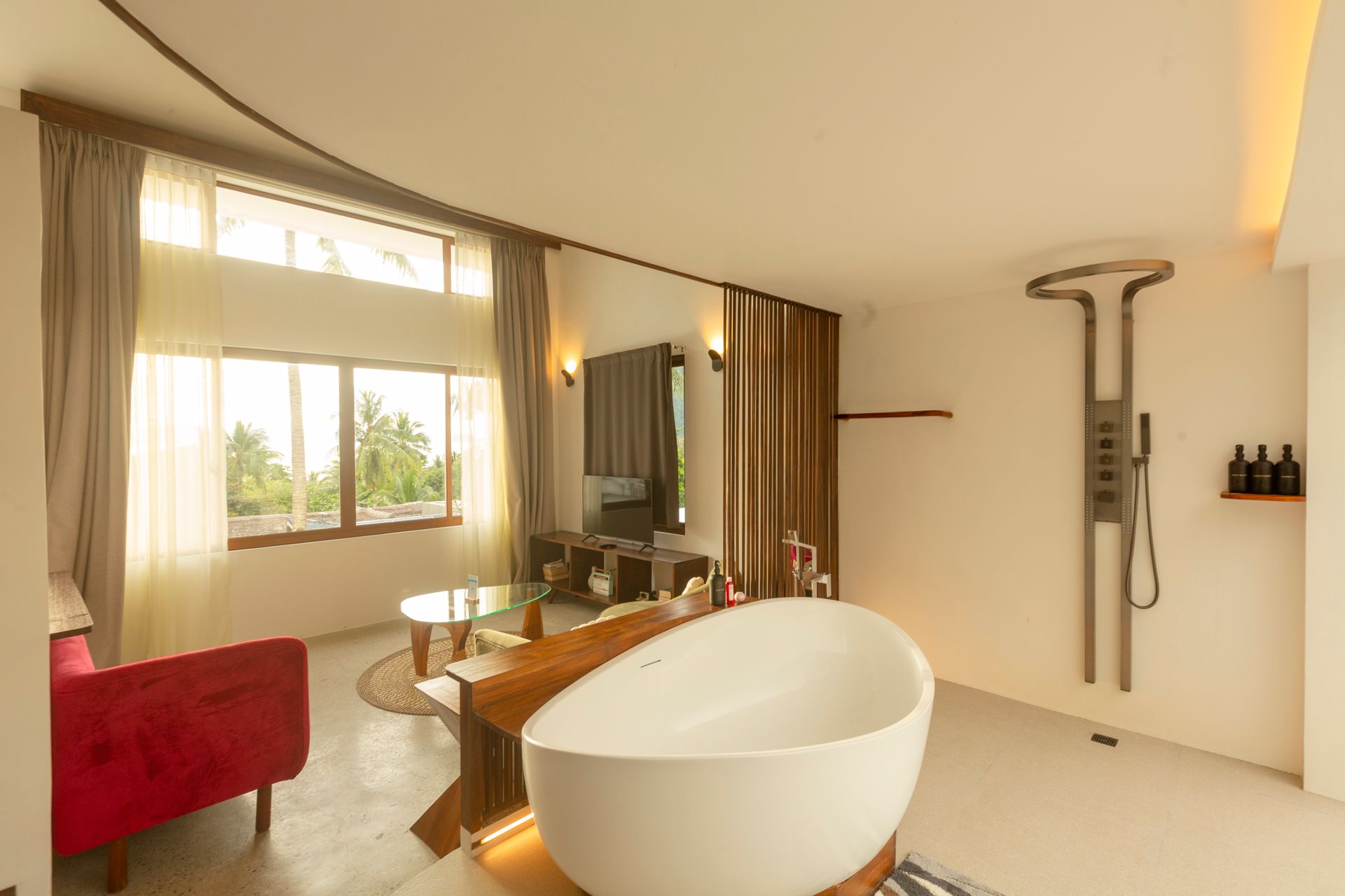 Modern hotel suite with a red sofa, a round table, and a white bathtub. Large windows offer a view.