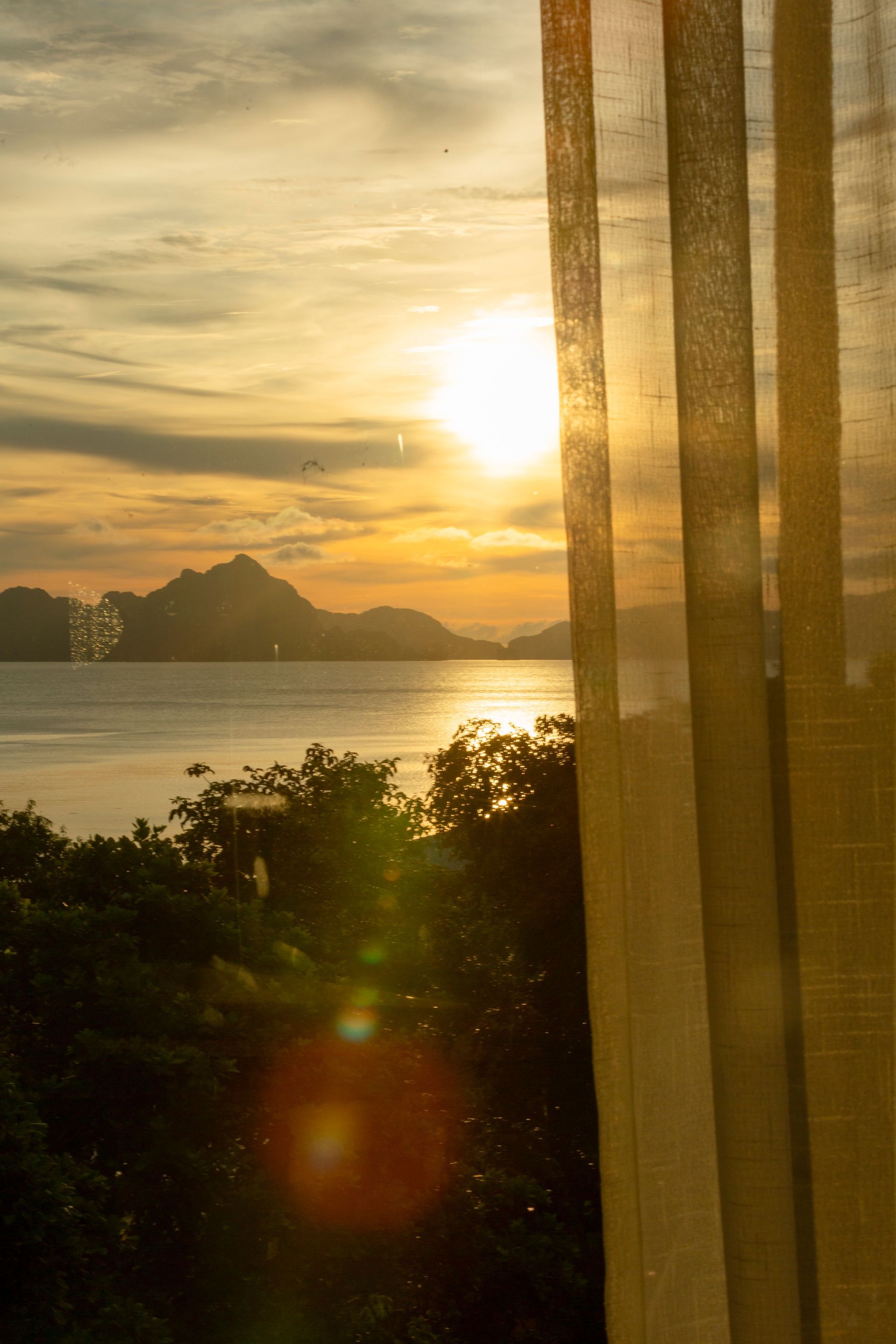 Sunset over water with silhouetted mountains and trees, viewed through sheer curtains.